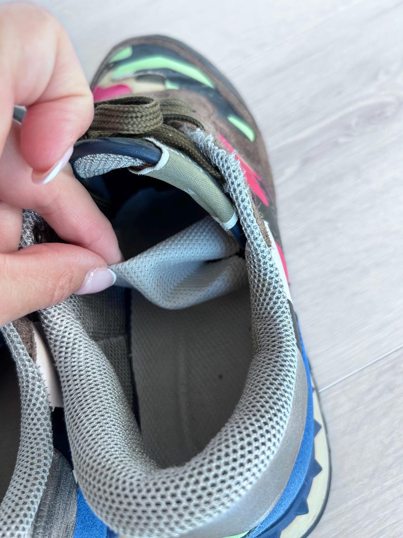 May include: Close-up of a multi-colored sneaker. The shoe features a camouflage pattern in shades of brown, green, and pink. The interior is light gray, and the laces are olive green. The sole has blue and gray accents.