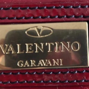 May include: Close-up of a glossy, deep red rectangular emblem with the brand name "VALENTINO GARAVANI" in gold lettering. The emblem is framed by a black stitched border.