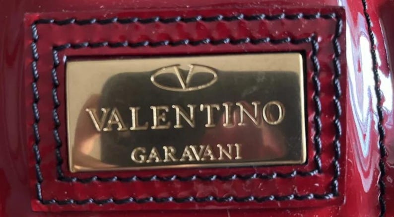 May include: Close-up of a glossy, deep red rectangular emblem with the brand name "VALENTINO GARAVANI" in gold lettering. The emblem is framed by a black stitched border.