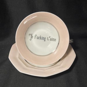 May include: A pale pink ceramic teacup and matching saucer set. The interior of the cup features a white base with the black text "Je fucking t'aime" printed in a serif font. The saucer has an octagonal shape with a smooth, light pink finish.