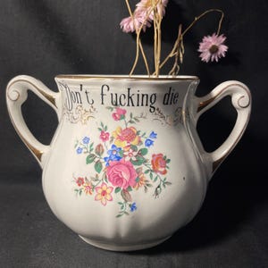 Può includere: Una zuccheriera in ceramica bianca con due manici e bordo dorato, caratterizzata da un colorato bouquet floreale e dal testo nero "Don't fucking die" stampato sul davanti. La ciotola contiene diversi fiori rosa essiccati con steli sottili.