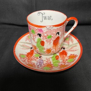 May include: A white ceramic teacup and saucer set featuring a hand-painted design of figures in traditional clothing, floral motifs, and an umbrella. The interior rim of the cup features the word Twat printed in a black, stylized serif font.