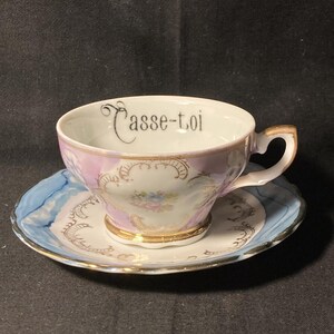 May include: A porcelain teacup and saucer set featuring a white base with light purple and blue accents. The cup interior displays the black text "Casse-toi" in a script font. Gold trim decorates the rim, handle, base, and floral patterns.