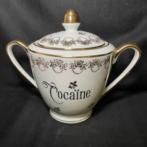 以下が含まれることがあります： 白いセラミックの砂糖入れ。蓋と2つの取っ手付き。金色のトリムと黒い筆記体で「Cocaine」の文字が特徴。ボウルは花柄とスクロール模様で装飾されています。