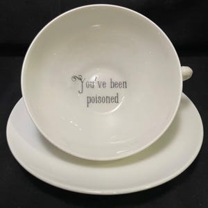 Könnte beinhalten: Weißes Tee-Set, bestehend aus Tasse und Untertasse. Die Tasse hat einen Henkel und die Worte "You've been poisoned" in Grau im Inneren. Die Untertasse ist rund und weiß.