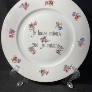 May include: A white ceramic plate with a gold-toned rim, decorated with small, colorful floral clusters in pink, blue, and orange. The center features the black text "Je baise mieux que je cuisine" surrounded by additional floral accents.