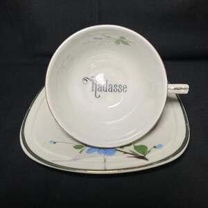 May include: A white ceramic teacup and matching saucer set featuring a silver rim and a delicate floral pattern with blue petals and green leaves. The interior of the teacup displays the word "Hadasse" in a stylized black cursive script font.