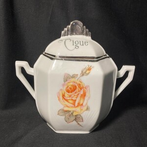 May include: A white porcelain sugar bowl with two handles and a matching lid. The lid features the word "Cigue" in script and a silver-toned finial. The body is decorated with a large yellow and orange rose illustration with green leaves.