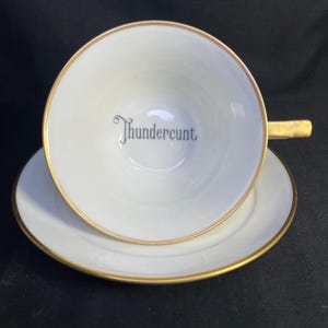 Peut inclure: Une tasse à thé et sa soucoupe en porcelaine blanche avec un bord et une anse dorés. Le fond intérieur de la tasse présente le mot Thundercunt imprimé dans une police serif sombre et stylisée. Le design est épuré, simple et centré.