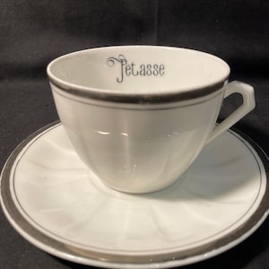 May include: A white porcelain teacup and saucer set featuring a silver rim and the word Petasse printed in black script on the interior of the cup. The cup has a faceted exterior surface and a small angular handle, while the saucer is round.