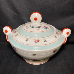 Può includere: Una zuppiera in ceramica bianca con coperchio. Il piatto presenta una fascia azzurra e motivi floreali, con la scritta "Valium" sul coperchio. Accenti rossi e bianchi sulle maniglie e sul coperchio.