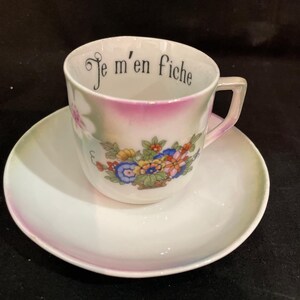 May include: A white porcelain teacup and saucer set featuring a colorful floral bouquet on the front and the black text Je m'en fiche printed along the inner rim. The cup has a handle and a soft iridescent finish with subtle pink and green accents.