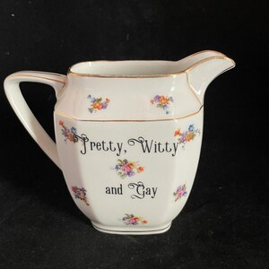 May include: A white porcelain creamer pitcher with a gold-trimmed rim and handle. The body features small, colorful floral clusters and the black printed text "Pretty, Witty and Gay" centered on the front. The pitcher has a faceted, angular shape.