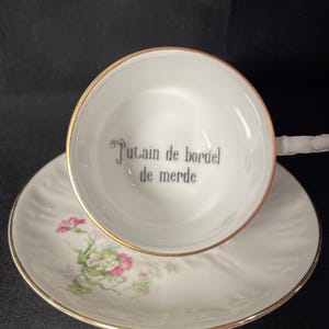 May include: A white porcelain teacup and saucer set with a gold rim. The interior of the cup features the black printed text "Putain de bordel de merde". The saucer is decorated with a small floral pattern of pink and green flowers on one side.