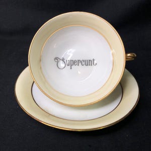 May include: A cream-colored teacup and saucer set with gold trim. The inside of the teacup features the word "Supercunt" in a stylized font. The saucer is white with a cream-colored border and gold trim.