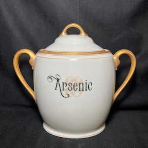 May include: White ceramic jar with a lid and two handles, featuring gold trim and handles. The word "Arsenic" is written in black script on the front, with a gold floral design beneath it. A decorative piece.