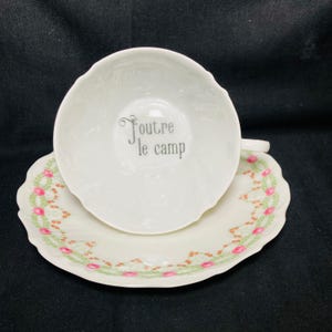 May include: White teacup and saucer set. The teacup has the phrase "Foutre le camp" printed in gray. The saucer is decorated with a floral pattern of pink and green. The set has a scalloped edge.