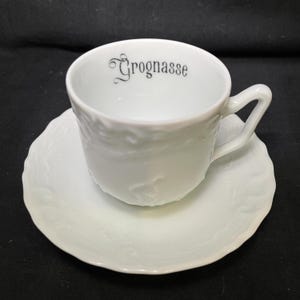 May include: A white porcelain teacup and matching saucer set featuring a delicate embossed floral pattern on the exterior. The interior rim of the cup displays the word Grognasse printed in a black, stylized serif font with a decorative initial G.
