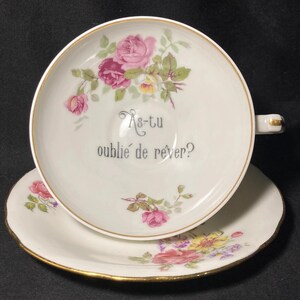 May include: A white porcelain teacup and saucer set featuring a gold rim and a floral pattern of pink and yellow roses. The interior of the cup displays the French text As-tu oublié de rêver? in a dark grey font, centered among the rose illustrations.