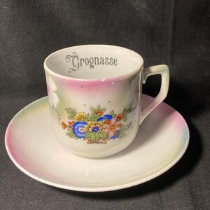May include: A white porcelain teacup and saucer set featuring a colorful floral bouquet on the front and the word Grognasse printed in black on the interior rim. The cup and saucer have a soft iridescent finish with subtle pink and green accents.