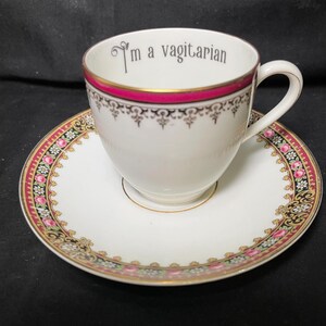 May include: A white porcelain teacup and saucer set featuring a pink and gold rim with a black floral pattern. The interior of the cup displays the text I'm a vagitarian in a dark grey font. The set has a classic handle and a matching floral border.