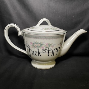 Peut inclure: Théière en céramique blanche avec couvercle, anse et bec verseur. La théière est ornée de motifs floraux et des mots "Fuck Off" en écriture noire. La poignée et le bord sont argentés.