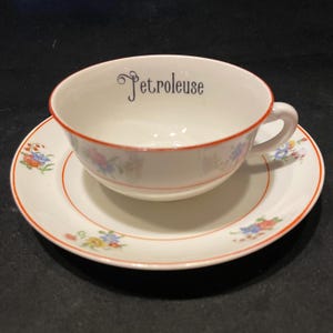 Op de afbeelding: Een witte keramische theekop en schotel met een dunne oranje rand en delicate bloemmotieven in blauw, geel en rood. De binnenkant van de kop toont het woord Petroleuse in zwarte schrijfletters, gecentreerd nabij de bovenste rand.