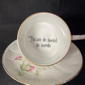 May include: A white porcelain teacup and saucer set with a gold rim. The interior of the cup features the black printed text "Putain de bordel de merde". The saucer is decorated with a small floral pattern in shades of pink and green.