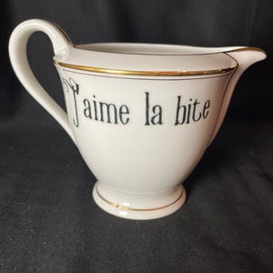May include: A white ceramic creamer pitcher featuring a gold-trimmed rim and base. The front of the pitcher displays the black text "J'aime la bite" in a stylized serif font. It includes a curved handle on the left side and a pouring spout.