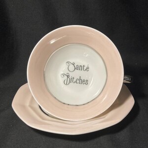 May include: A pale pink teacup and matching saucer set. The interior of the white teacup features the black text "Santé Bitches" written in a decorative script font. The teacup has a smooth, rounded rim and a small handle on the right side.