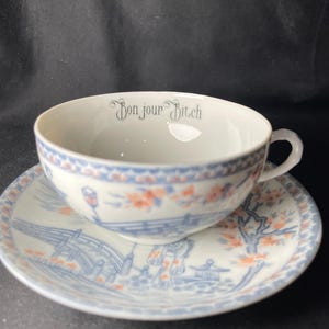 May include: A white porcelain teacup and saucer set featuring a blue and orange landscape pattern with a bridge and trees. The interior rim of the teacup displays the text "Bonjour Bitch" in a dark grey, decorative serif font. The set has a handle.