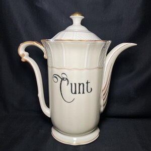 May include: Cream ceramic coffee pot with a lid and curved handle. The pot has gold trim and the word "Cunt" in black script on the front. The pot is set against a black background.