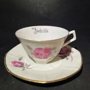 May include: A white porcelain teacup and saucer set featuring a delicate pink rose floral pattern and gold trim. The interior rim of the teacup displays the black text Boobzilla in a decorative script font, with a matching rose design on the saucer.