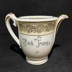 May include: Cream-colored ceramic creamer with gold trim and handle. The creamer features a decorative band with a paisley design in gold and the text "Fuck Trump" in black script.