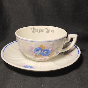 May include: A white porcelain teacup and saucer set featuring a blue floral pattern and a thin blue rim. The interior of the teacup displays the black text Bon jour Bitch in a decorative script font. The cup has a geometric handle design.