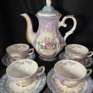 May include: A porcelain tea set featuring a teapot and four teacups with matching saucers. The pieces are light purple and blue with gold trim, floral accents, and the text "Fuck Off" printed in black script on the teapot and inside each cup.