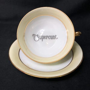 May include: A porcelain teacup and saucer set featuring a cream-colored rim and a gold-toned border. The interior of the white teacup displays the word Supercunt printed in a dark gray, stylized cursive font. The handle is also gold-toned.