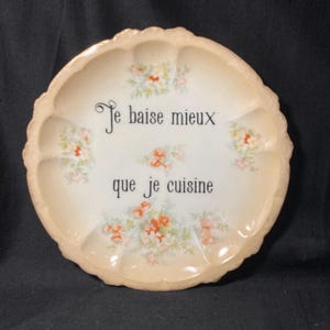 May include: A round, cream-colored ceramic plate with a scalloped edge and a gold-toned rim. The plate features floral clusters in shades of orange, pink, and green, with the black text "Je baise mieux que je cuisine" printed in the center.