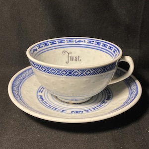 May include: A white ceramic teacup and saucer set featuring a textured dimpled surface and blue geometric border patterns. The interior of the teacup displays the word Twat in a black script font, centered against the white background.
