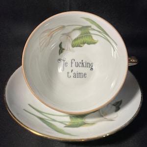 May include: A white porcelain teacup and saucer set featuring a gold-trimmed rim and a delicate green and white calla lily floral pattern. The interior of the cup displays the black printed text "Je fucking t'aime" in a classic serif font.