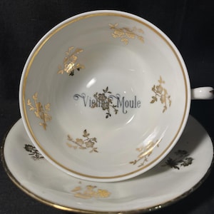 May include: A white porcelain teacup and saucer set featuring a gold rim and delicate gold floral patterns. The interior of the cup displays the text "Vieille Moule" in black script, surrounded by a small gold floral design at the center base.