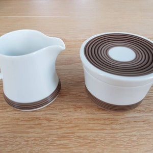 Op de afbeelding: Een witte keramische roomkan en suikerpot set met bruine concentrische cirkelontwerpen.