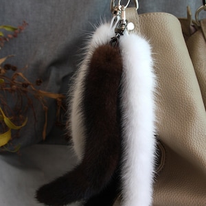 Può includere: Un portachiavi con tre code di pelliccia soffice, nere, marroni e bianche. Sono attaccate a un fermaglio in metallo argentato, appese vicino a una borsa in pelle beige. La pelliccia appare morbida e lussuosa.