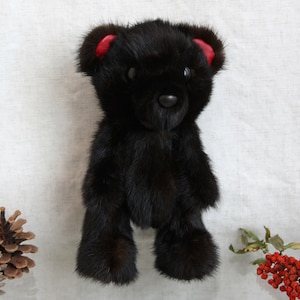 May include: A black fur teddy bear with red ears. The bear is standing on a white fabric background.