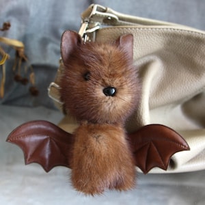 May include: A brown plush bat keychain with leather wings. The bat has a fluffy body, small ears, and a black bead for an eye. The keychain is attached to a beige handbag, suggesting it's a decorative accessory.