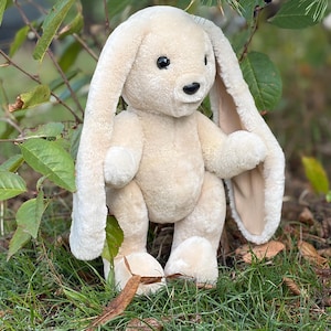 Pode incluir: Um peluche de coelho com pelo macio de cor creme, orelhas longas e caídas, e olhos de plástico escuro. O coelho tem uma cabeça arredondada, um pequeno nariz preto e uma boca cosida simples, de pé com um corpo redondo e membros curtos.