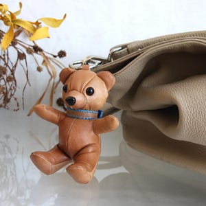 Op de afbeelding: Een bruine leren teddybeer sleutelhanger met gestikte panelen, zwarte kraaloogjes, een zwarte neus en een blauwe stoffen halsband. De beer is bevestigd aan een zilveren metalen ring en bevindt zich naast een beige leren handtas.