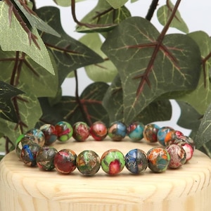 Puede incluir: Un brazalete de cuentas multicolores y estampadas. Las cuentas son predominantemente rojas, verdes, azules y naranjas. El brazalete se exhibe sobre un pequeño soporte de madera, con hojas verdes en el fondo.