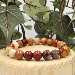 Puede incluir: Un brazalete de cuentas con tonos tierra, incluyendo piedras rojas, marrones y crema. Se exhibe sobre un pequeño soporte redondo de madera, con follaje verde en el fondo.