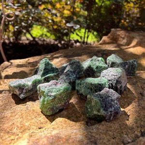 May include: A cluster of raw zoisite crystals with ruby flecks. The stones show a mix of green, black, and red tones, set on a textured, light brown surface. Natural light accentuates the crystals' diverse textures and colours.